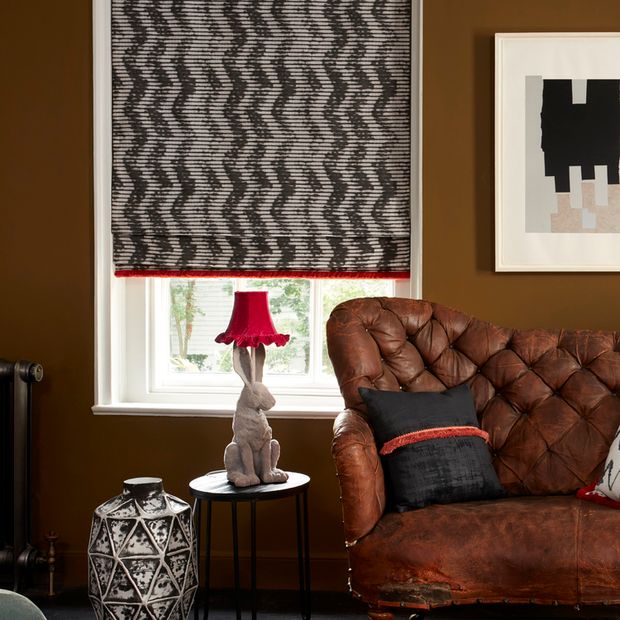 Roman blind with a zig zag pattern in black and white is fitted to a window in a living room with a brown leather sofa, the sidetable next it has a lamp shaped like a rabbit with a red coloured pendant