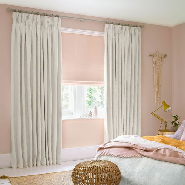 White coloured curtains which are matched with pink roman blinds are fitted to a rectangular shaped window in a pink decorated bedroom
