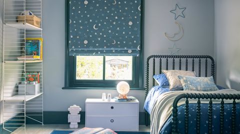 A child's bedroom with a blue roman blind featuring stars and moons. A star and moon decoration hangs on the right above the bed.