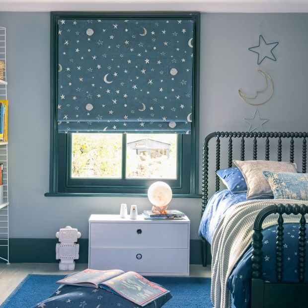 A child's bedroom with a blue roman blind featuring stars and moons. A star and moon decoration hangs on the right above the bed.