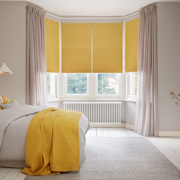 Mustard yellow blackout Pleated blinds dressed on windows in bedroom. Light pink Voile curtains are hanging over the blinds. Matching mustard and grey cushions have been placed on the bed.