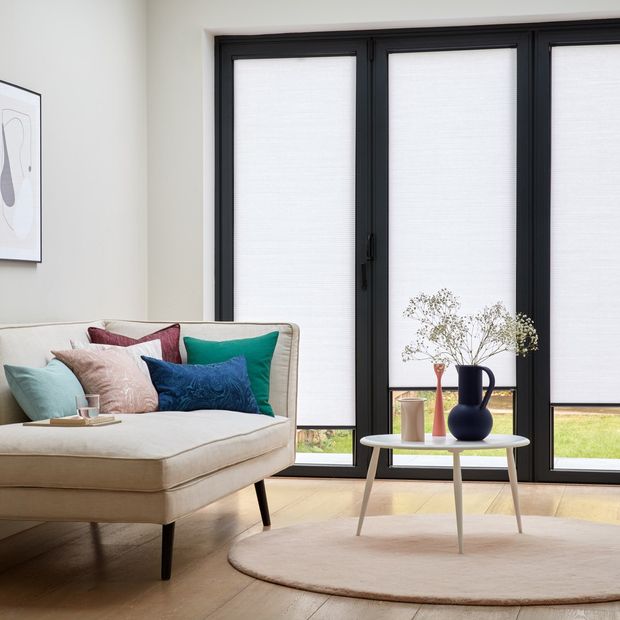White Pleated blinds dressed on bi-fold doors of living room. Red, cream, green and blue cushions have been placed on cream corner sofa bed in room.