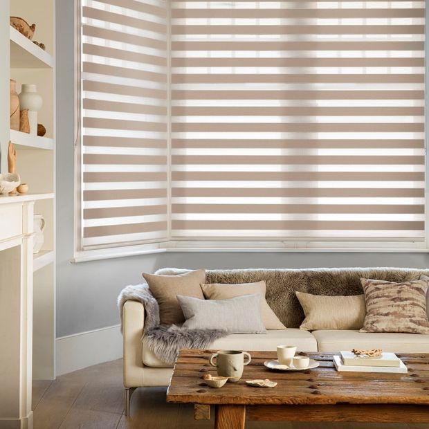 prism taupe day and night blinds in a bay window in a living room with a wooden table, furnished cream sofa and cream shelving