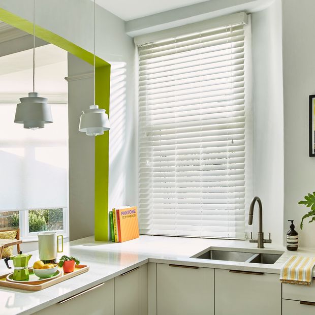 Mirage White Maple blind in a white kitchen with lime features. There is an attached breakfast bar with a tray of breakfast food and drinks on it. There is a sink with a plant next to it in the right side and a wooden table with chairs on the left side.