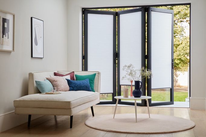 white nook pleated blinds fitted on bifold door windows which are half open from a sitting area into the garden