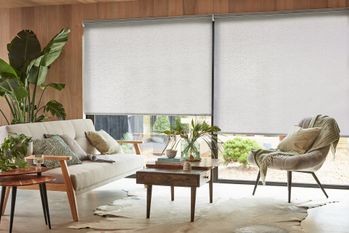 Living room with wooden walls and celing, wide patio doors dressed in pale grey roller blinds, halfway and three quarters lowered, pale animal skin rug under contemporary cream and wood sofa and chair, and wooden coffee table.