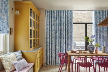Dining room with wooden table and wooden chairs painted dusky pink, wooden dresser on left with blue and white marble top, ming-style vase on table and curtains with pen and ink style small sketches in blue on a white background, foreground partial window dressed in matching Roman blind.