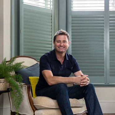 George clarke sat on a cream chair in a living room that has teal coloured shutters