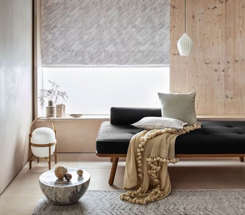 The room features a window partially covered with a soft grey Roman blind. In front of the window and to the left is a long black couch that has a beige throw draped across it. This room also has an exposed wooden wall and minimal accessories to the right of the chair
