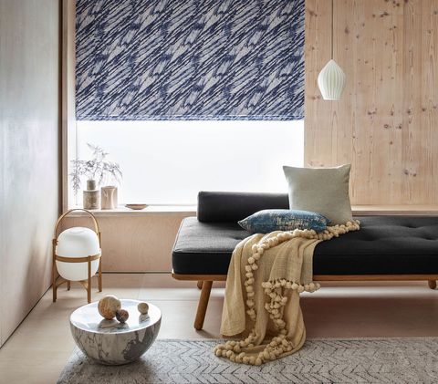 A window partially covered with blue patterned roman blinds in a siting room, matching cushions and cream shawl have been placed on a soft top table and room is decorated with flower vases