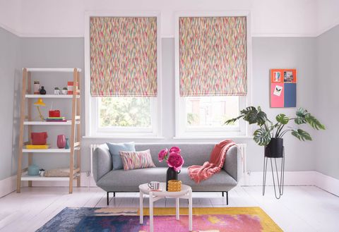 This grey and white room features a grey couch with cushions and a throw. Multi-coloured Roman blinds hang half way down the white windows