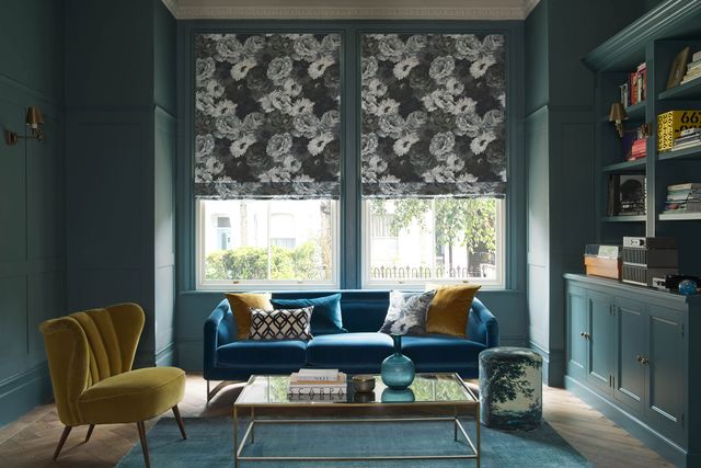 A blue couch rests in front of a window that is covered by a black floral Roman blind. The room is painted a deep forest green