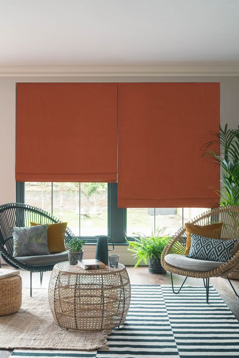 Orange  roman blinds in a garden room. Room is decorated with wicker chairs and table. Blue diamond patterned cushion, mustard chushions and leafy printed cushion have been placed on chairs.