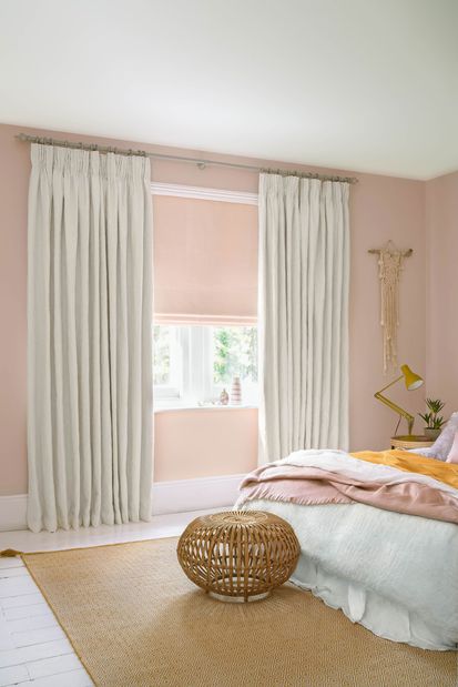 White coloured curtains which are matched with pink roman blinds are fitted to a rectangular shaped window in a pink decorated bedroom