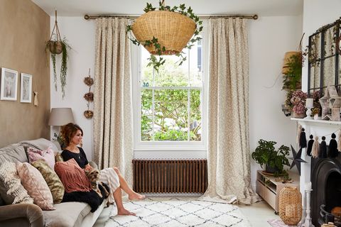 A woman sat in a white and honey-coloured living room with a single sash windows dressed with cream and white patterned curtains