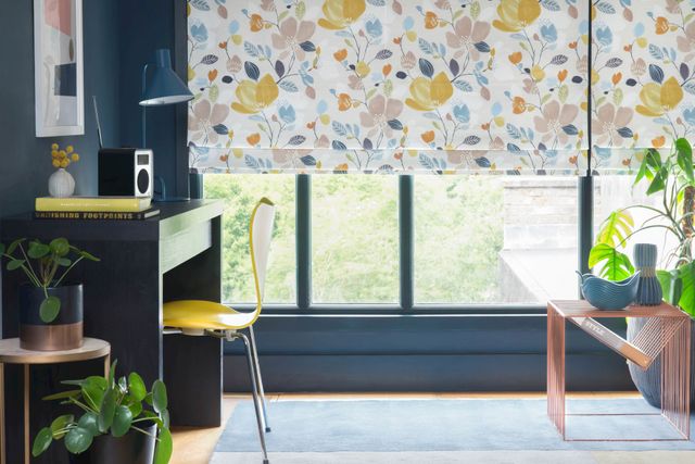 A multi-coloured floral Roman blind hangs neatly in front of a window on a petrol blue wall. Plants are dotted around the room and there is a desk to the left of the window. 