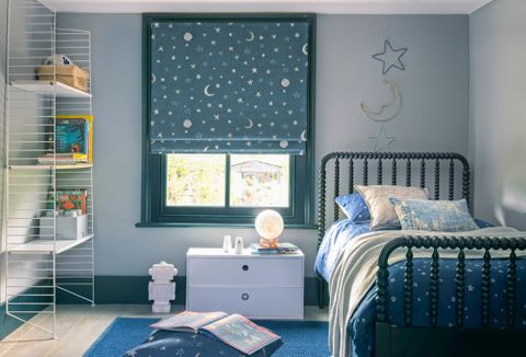 A child's bedroom with a blue roman blind featuring stars and moons. A star and moon decoration hangs on the right above the bed.