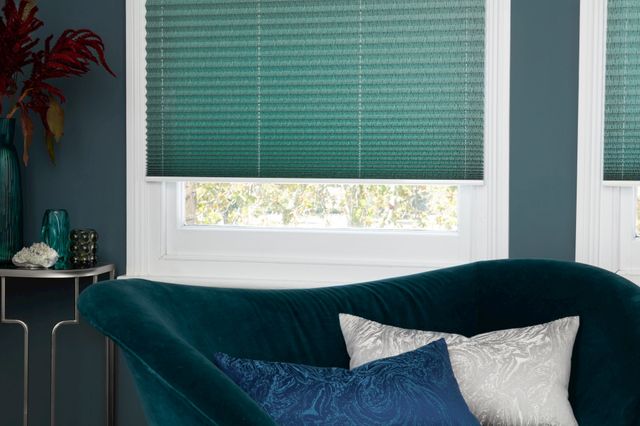 Green Pleated blinds from House Beautiful range dressed on windows in living room. Green velvet sofa has been placed by window.