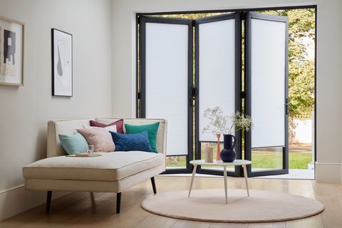 White Pleated blinds dressed on bifold doors. Block colour cushions on cream corner sofa bed, Doors are open.