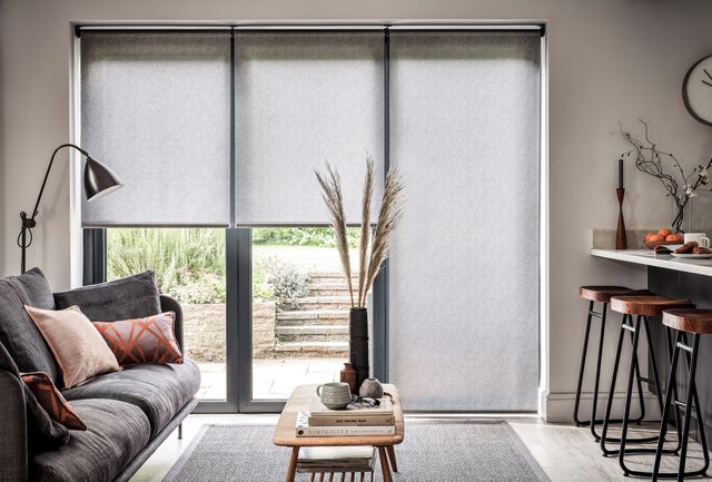 Grey roller blinds in a living room with a grey sofa on the left a table in the centre and a breakfast bar with stools on the right.