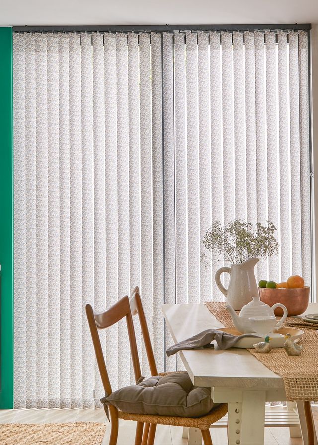 Vertical white blinds in a dining room with a wooden table and chairs surrounding the table. There is a vase and a bowl of fruit on the table. 