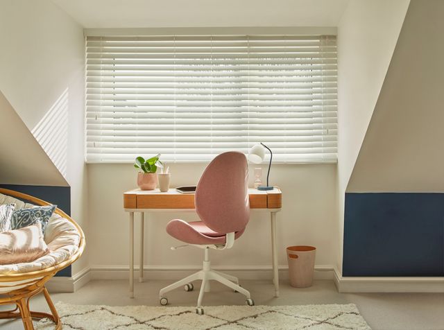 Wood venetian Illusions Faux Wood Matt White Blinds at a window with a desk and table in front of it and a comfy chair on the left side. 