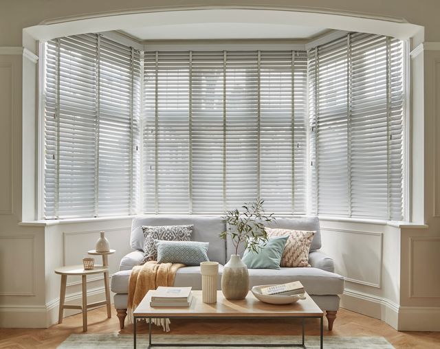 Wood Venetian Ellwood Silver Grey Blinds in a living room at a bay window. A sofa and a coffee table sit in front of the window. 