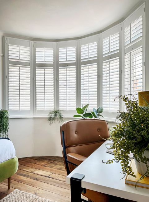 Pure white full height bay window shutter in a bedroom with wooden floor, and a desk with a plant on it and an office chair