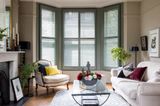 dark green full height shutters in a living room two white sofas glass table and black fire place surrounded by cream walls