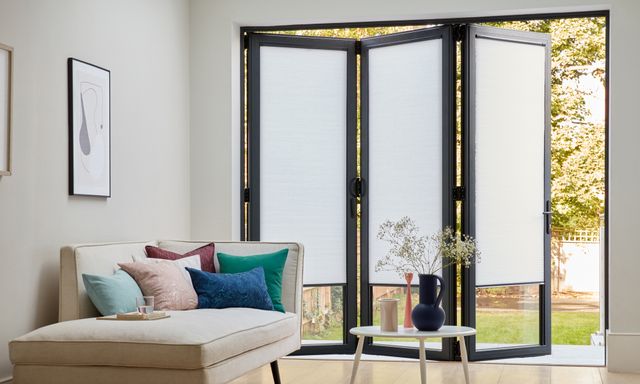 white nook pleated blinds fitted on bifold door windows which are partially open from a sitting area into the garden 