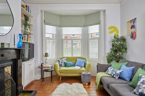 A living room with a wood floor, feature fireplace, sheepskin rug and a small chartreuse velvet sofa in front of a bay window dressed with white café style shutters to midway, pale green Roman blinds partially lowered, latered over white sheer Roller blinds.