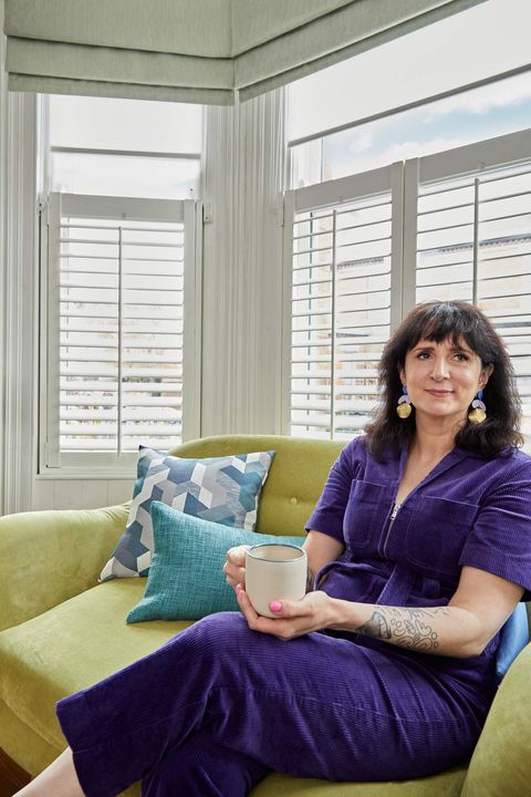 A woman sits on a chartreuse velvet two seater sofa in a living room with a wood floor, in front of a bay window dressed with white café style shutters to midway, pale green Roman blinds partially lowered, latered over white sheer Roller blinds.