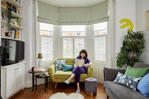 A woman sits on a chartreuse velvet two seater sofa in a living room with a wood floor, in front of a bay window dressed with white café style shutters to midway, pale green Roman blinds partially lowered, latered over white sheer Roller blinds.