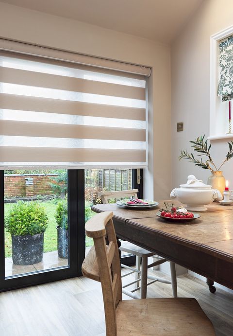 Cream living room with small window dressed in Roman blind in white with green foliage pattern and main windows and door to rear with beige day and night blind, lowered halfway, tilted partially open, rustic wooden dining table and chairs, red candles.